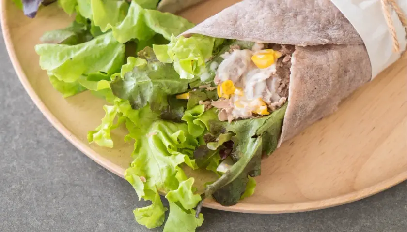 Pyszna tortilla z tuńczykiem, świeżą sałatą i kukurydzą, zawinięta w papier i przewiązana sznurkiem.