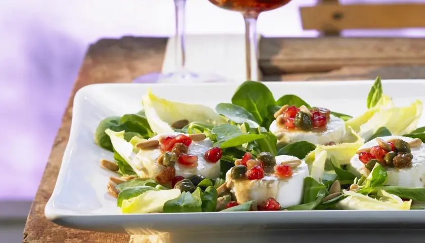 Ein frischer Salat mit Ziegenkäse, garniert mit Granatapfelkernen und Pinienkernen, serviert auf einem weißen Teller.