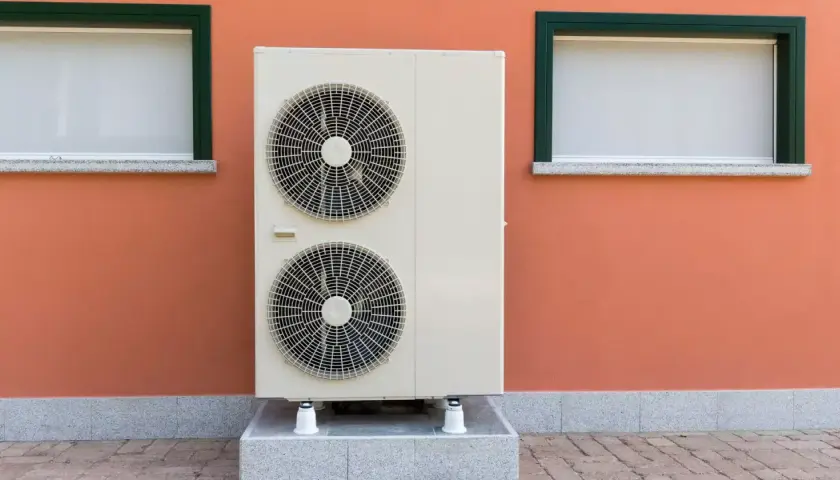 Unidad exterior de bomba de calor con dos ventiladores, instalada junto a una pared naranja con dos ventanas.