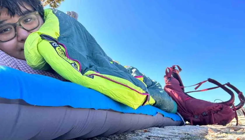 A person rests on a sleeping pad under a bright blue sky, bundled in a Nemo Disco 15 weight sleeping bag. A red backpack sits nearby.