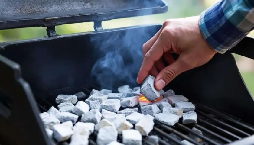 Une main allume des briquettes de charbon dans un barbecue, de la fumée s'échappe. La question est : combien de temps pour allumer un barbecue ?