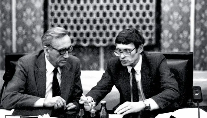Dwóch mężczyzn w garniturach przy stole, dyskutujących o historii politycznej Polski. Antoni Dudek analizuje dokumenty.