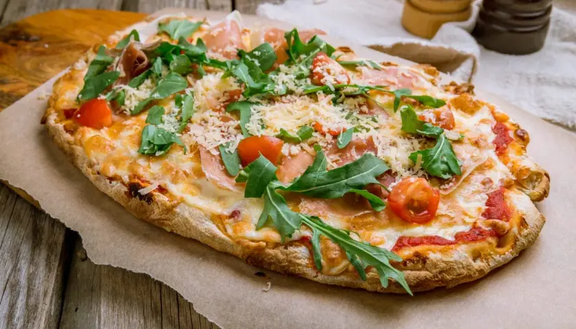 Pyszna pinsa romana z rukolą, pomidorkami i szynką parmeńską, posypana parmezanem, na drewnianym stole.