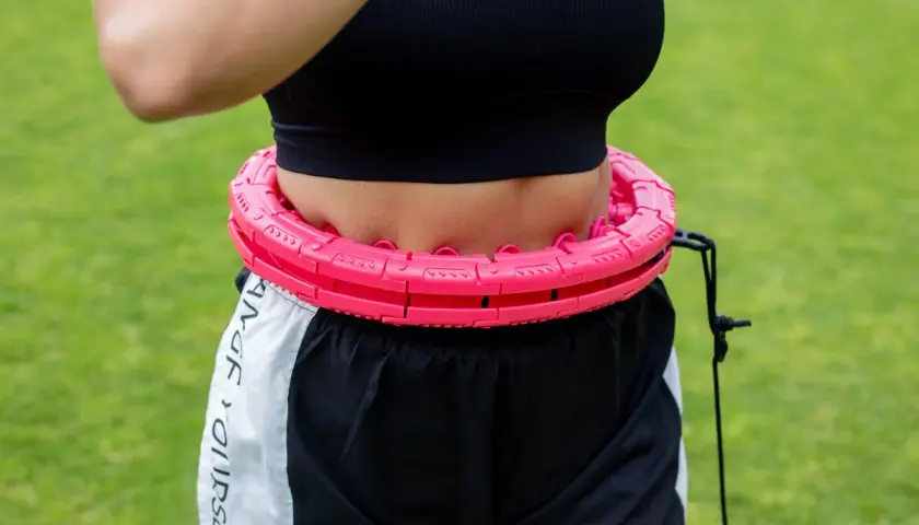 Kobieta w sportowym topie i szortach wykonuje ćwiczenia z hula hop na trawie. Różowe, segmentowe hula hop z obciążnikiem.