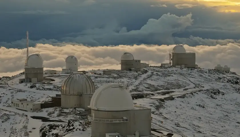 Obserwatoria astronomiczne na ośnieżonym szczycie, otoczone morzem chmur. W oddali widać złote światło zachodzącego słońca, a na pierwszym planie "la silla" - jedno z kopułowych budynków.