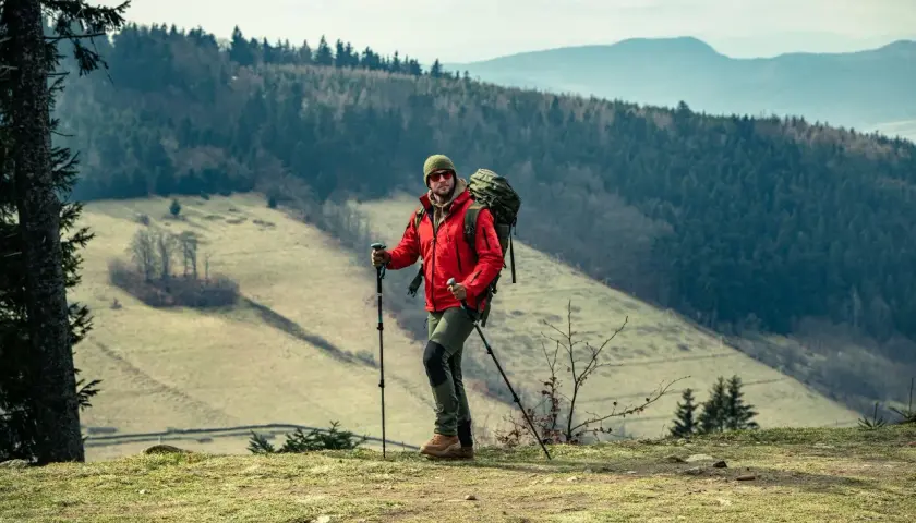 Mężczyzna w czerwonej kurtce i z plecakiem uprawia nordic walking na tle górskiego krajobrazu.