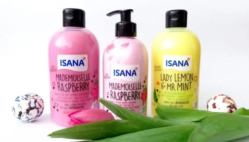 Trzy butelki ISANA: "Mademoiselle Raspberry" (różowa, z pompką i bez) i "Lady Lemon & Mr. Mint" (żółta). Obok kwiaty i ozdobne kule.