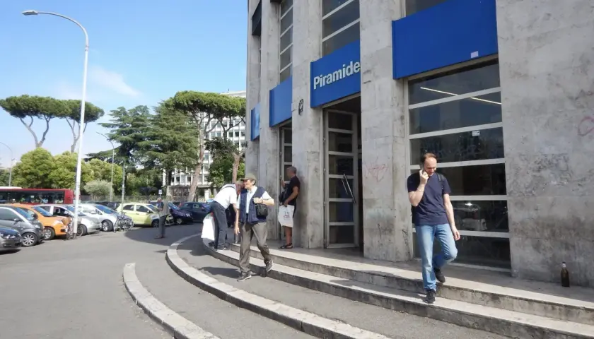 Uscita della metro Piramide a Roma Ostiense. Persone entrano ed escono dall'edificio, mentre auto sono parcheggiate nelle vicinanze.