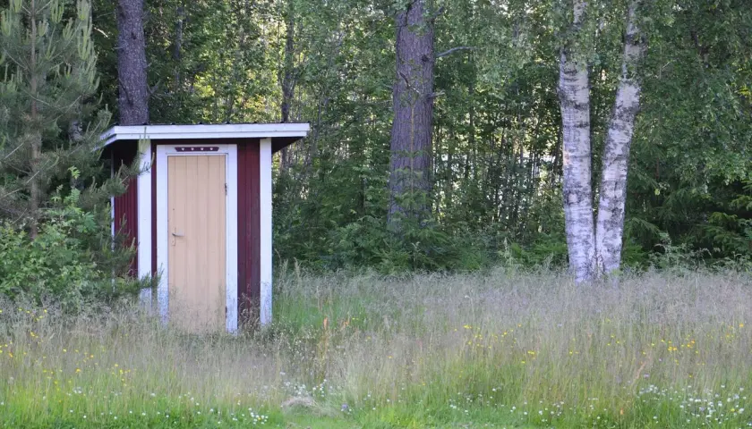 Rött utedass med gul dörr, omgivet av högt gräs och träd. Här gäller utedass regler för en trevlig vistelse.