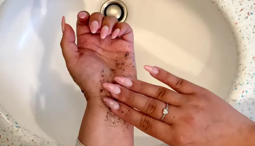 Exfoliante de café en la mano para quitar el autobronceador. Uñas largas y rosadas, anillo de corazón.