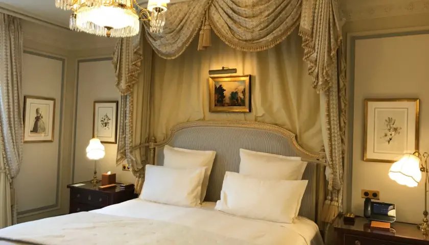 Elegancka sypialnia w hotelu Ritz w Paryżu, z baldachimem nad łóżkiem, kryształowym żyrandolem i zabytkowymi obrazami.
