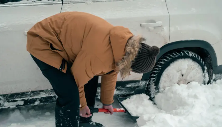 Osoba w zimowej kurtce odśnieża samochód uwięziony w zaspie śnieżnej.