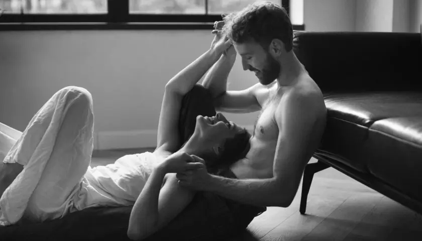 Para przytulająca się na podłodze. Kobieta śmieje się, opierając głowę na jego kolanach. Scena sugeruje intymność, ale nie jest to seksualizacja kobiet.