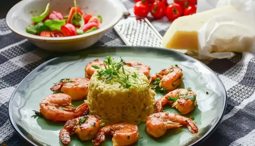 Krewetki z mlekiem kokosowym i curry na risotto, udekorowane ziołami. Obok miseczka z pomidorkami i bazylią, ser i tarka.