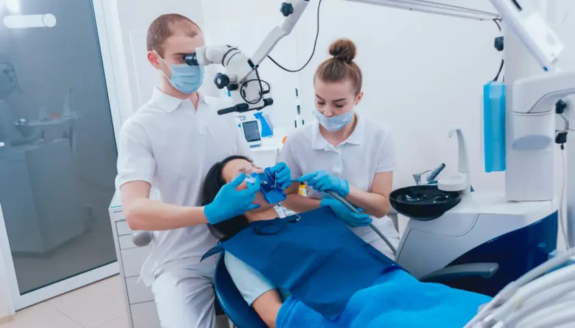 Dentysta pod mikroskopem bada ząb pacjentki, która leży na fotelu. Asystentka przygotowuje się do zabiegu, by zapobiec zatruciu zęba.
