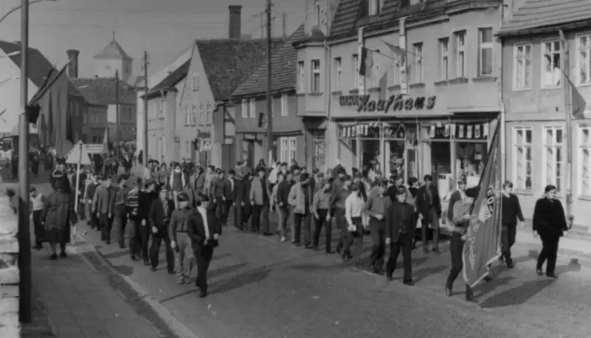 Demonstration am 7. Oktober, Tag der Republik DDR. Menschen marschieren durch eine Straße mit Geschäften, Fahnen wehen.