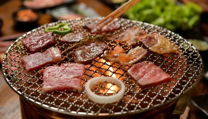 Enjoying delicious yakiniku at home. Slices of raw beef sizzle on a grill over glowing coals, with green peppers and an onion ring adding color.