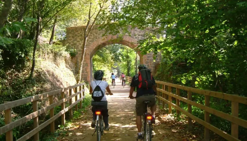 Ciclistas disfrutan de la vía verde Olot Girona, pedaleando bajo un antiguo arco de piedra rodeado de exuberante vegetación.