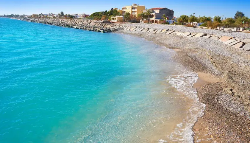 Aguas turquesas bañan la playa almenara, con un rompeolas de rocas y edificios al fondo.