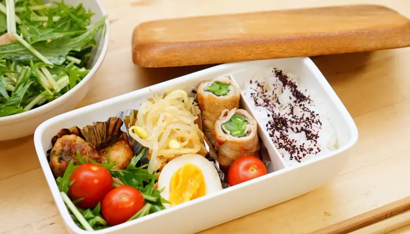 A delicious rice bento box with pork rolls, chicken meatballs, a hard-boiled egg, cherry tomatoes, and a side salad.