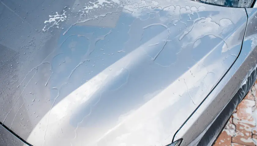 Krople wody spływają po masce srebrnego auta, tworząc smugi. Czy po woskowaniu płukać auto? Tak, aby uzyskać idealny połysk.