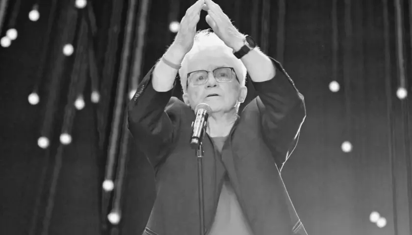 Starsza pani z białymi włosami, w okularach, z rękami uniesionymi nad głową, przed mikrofonem. To może być Jolka podczas występu.