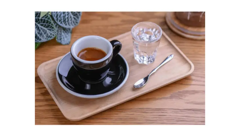 Czarna filiżanka espresso na drewnianej tacy, obok szklanka wody i łyżeczka. Idealnie pokazuje, jak podawać espresso.