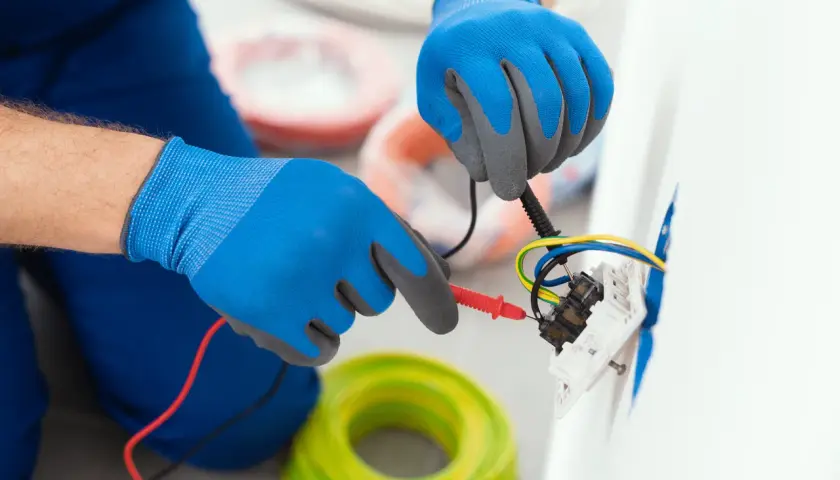 Elektryk w niebieskich rękawicach sprawdza połączenia przewodów. W tle widać rolki kabli. To instrukcja, jak podłączyć gniazdko bez uziemienia.