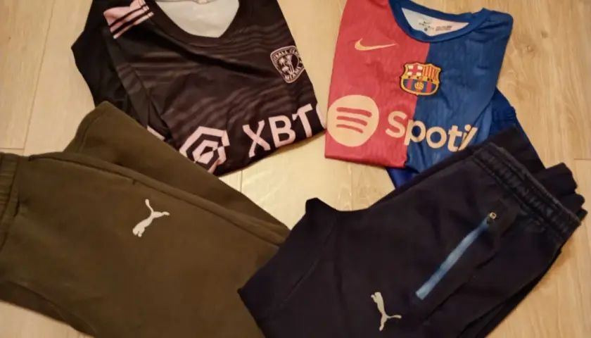 Dwa komplety odzieży piłkarskiej dla chłopca: czarna koszulka z różowymi pasami i herbem, niebiesko-czerwona koszulka FC Barcelony, brązowe i granatowe spodnie piłkarskie chłopięce z logo Pumy.