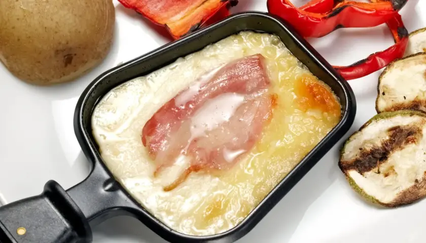 Un poêlon de raclette avec fromage fondu et lardons, accompagné d'une pomme de terre, de poivrons rouges grillés et de courgettes. Une recette de sauce raclette facile et gourmande.