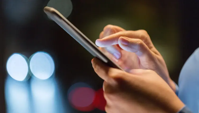 Dłoń trzyma smartfon, palec wskazujący dotyka ekranu, być może w trakcie odzyskiwania zdjęć z telefonu. Rozmyte światła w tle sugerują noc.