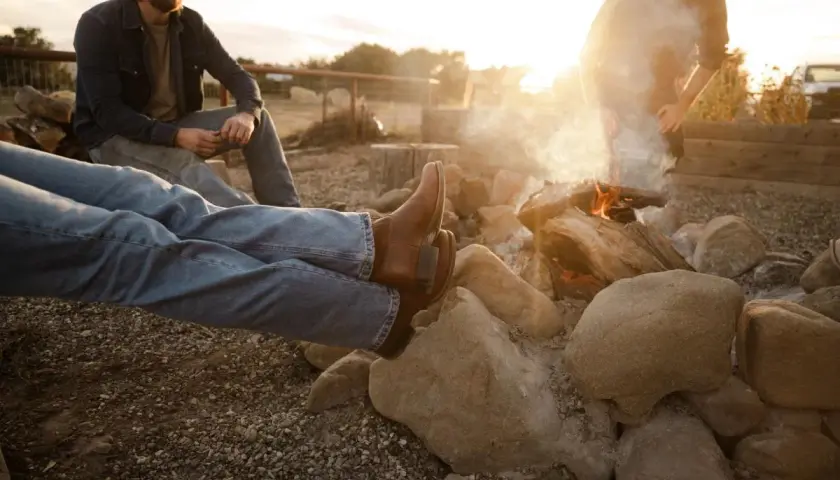 Two men relax by a campfire. One wears brown boots, and the other, a cap. The sun sets, casting a warm glow.