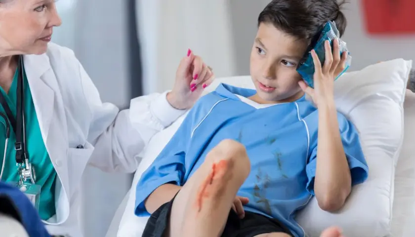 Niño con golpe en la cabeza, recibe atención médica. Tiene una compresa fría en la sien y una herida en la rodilla.