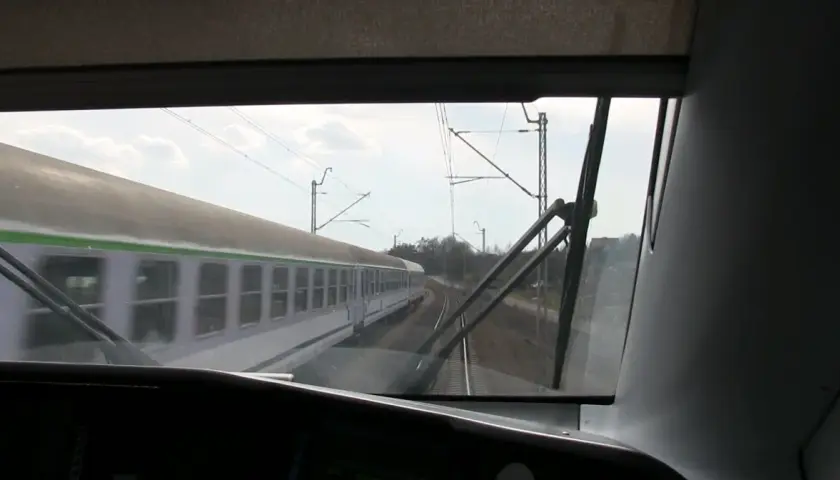 Widok z kabiny maszynisty na mijający pociąg. Widać tory, sieć trakcyjną i niebo z chmurami.