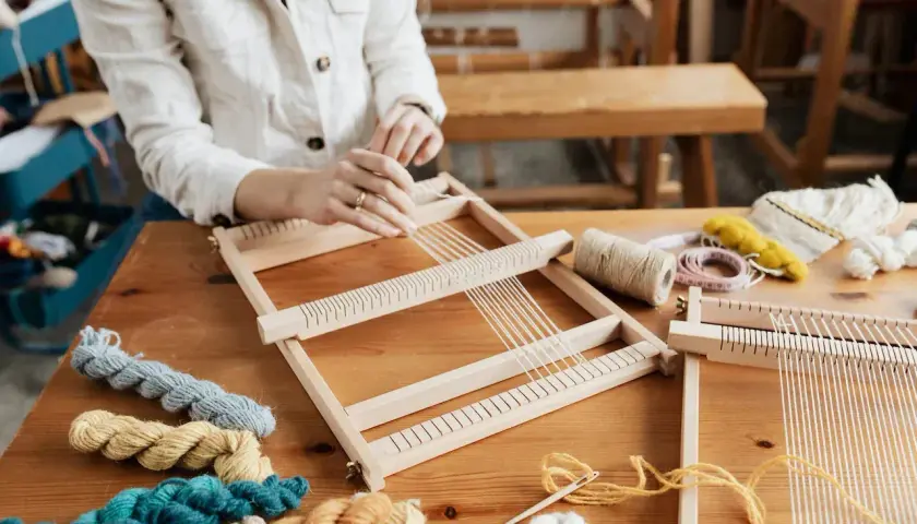 Hände weben auf einem Holzrahmen. Bunte Wollknäuel und Werkzeuge liegen bereit. Tolle Ideen für Erwachsene zum Weben.