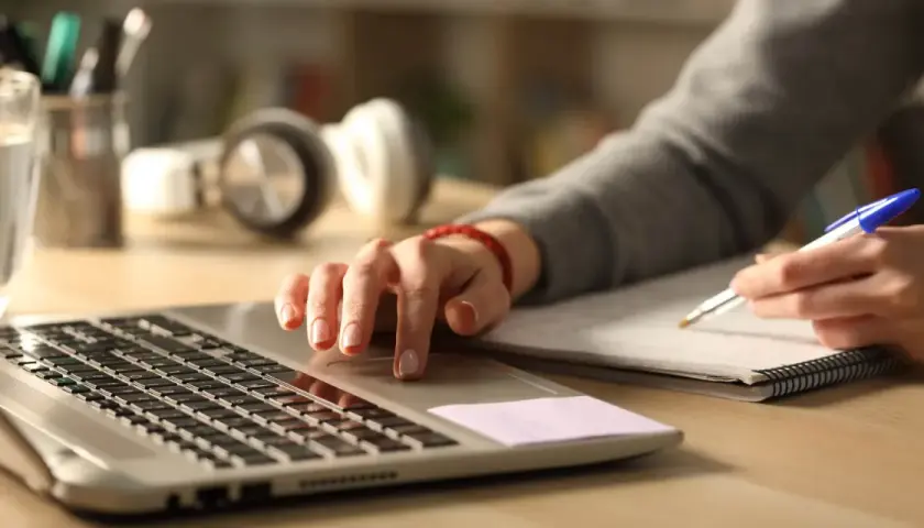 Personens händer arbetar vid en laptop och skriver i en anteckningsbok. Perfekt för kreativt skrivande distans.