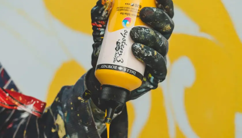Mano con guanti sporchi di vernice che stringe un flacone di giallo brillante, pronto a creare un'opera d'arte. Il colore viola è assente, ma l'energia creativa è palpabile.