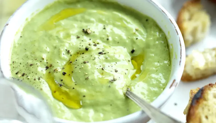 A bowl of creamy green basil pesto with olive oil and cracked pepper, served with toasted bread.