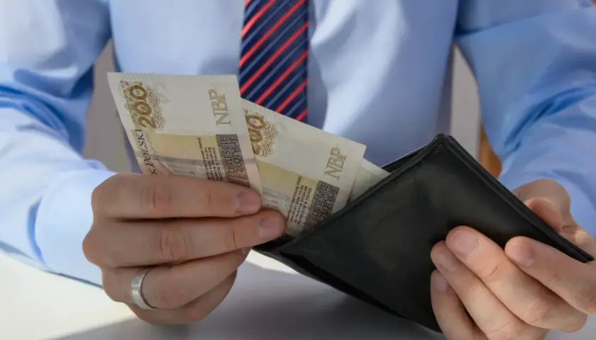 Mężczyzna w koszuli i krawacie wkłada banknoty 200 zł do portfela. Może to być wypłata, premia lub odprawa pośmiertna.