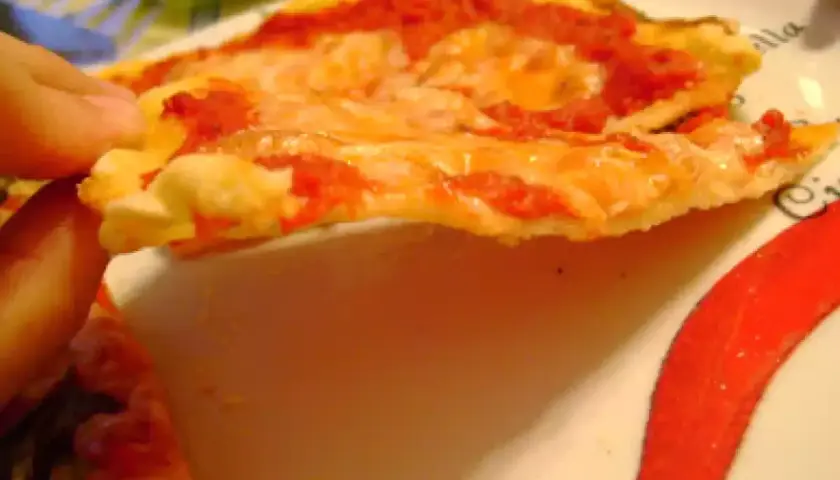 Una mano sostiene una rebanada de pizza con queso derretido y salsa de tomate, sobre un plato decorado con pimientos rojos. La masa, hecha con semola de trigo, luce dorada y crujiente.