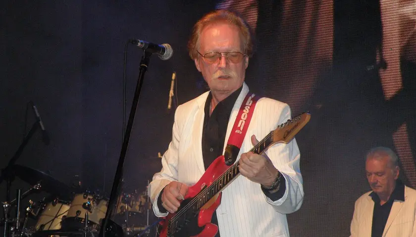 Muzyk w białej marynarce gra na czerwonej gitarze. Na scenie widać też perkusję i klawisze.