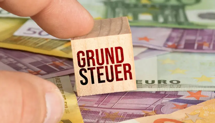 Ein Finger hält einen Holzstein mit der Aufschrift "GRUNDSTEUER" über Euro-Banknoten. Der **Grundsteuer Messbetrag** ist hier visuell dargestellt.