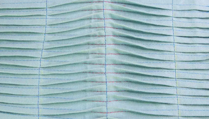 Close-up of light blue fabric with rows of pintucks, creating a textured, pleated effect. Red and blue threads highlight the stitching.
