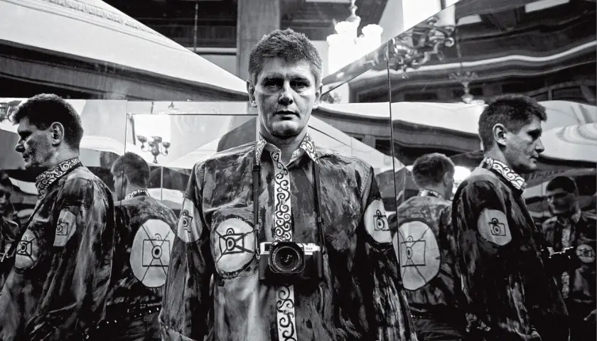 Krzysztof Miller w lustrzanym odbiciu, z aparatem na szyi, w koszuli z motywem oka i aparatu.