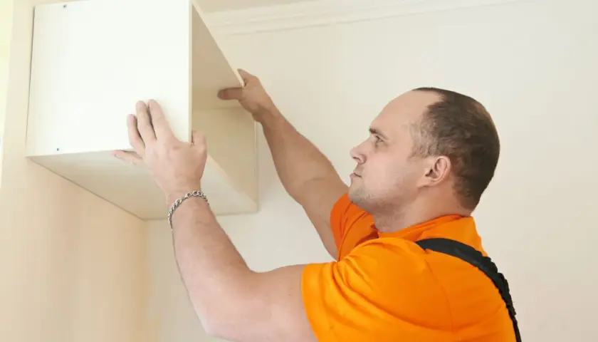 Un homme en orange est en train de fixer un meuble lourd au mur. Il positionne soigneusement l'étagère avant de la visser.
