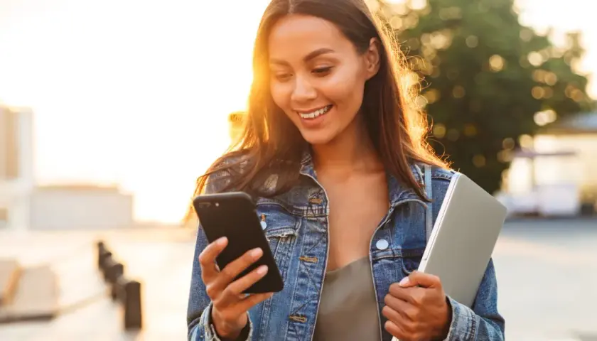 Uśmiechnięta dziewczyna w dżinsowej kurtce czyta na telefonie dwuznaczne teksty do dziewczyny, trzymając laptopa.