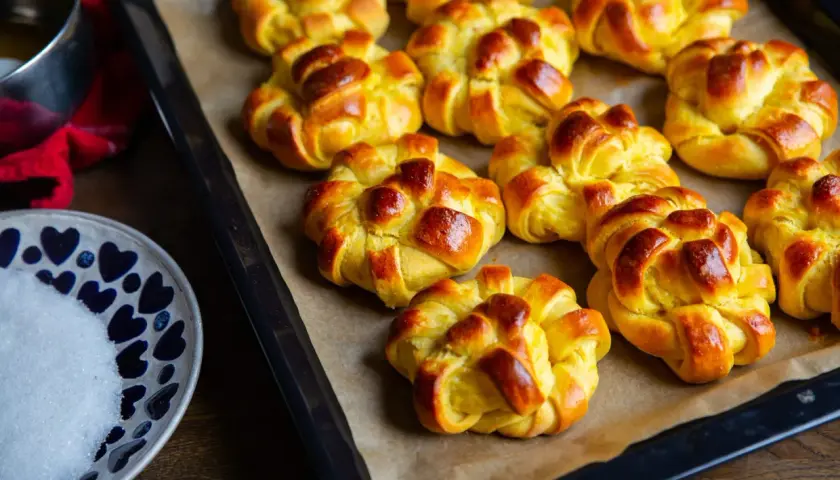Nybakade zeina saffransbullar på en plåt, gyllene och inbjudande. En skål med socker och en röd duk anas i bakgrunden.