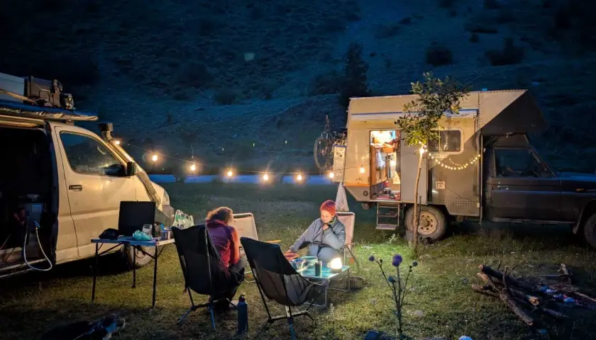 Soirée paisible autour d'un feu de camp, deux personnes assises près d'un **van aménagé électrique** illuminé par des guirlandes lumineuses.