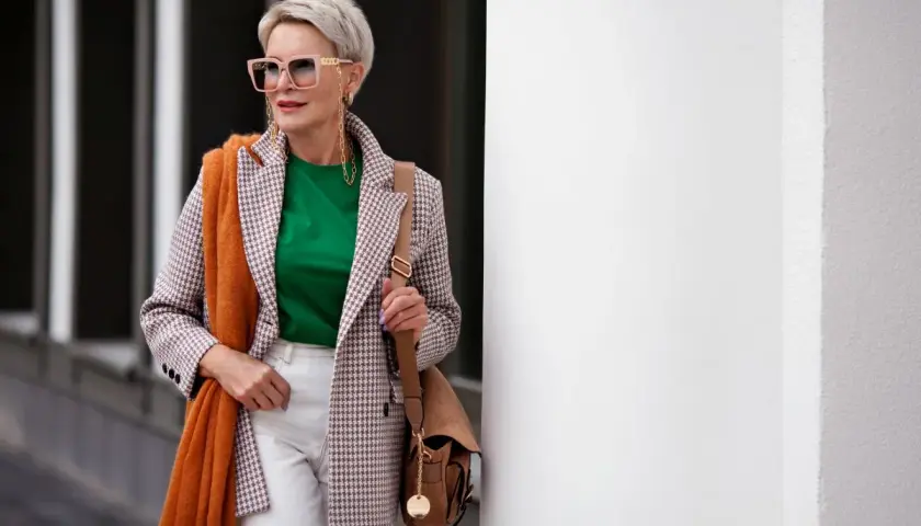 Elegancka kobieta po 60. w stylowej marynarce w kratkę, zielonej bluzce i białych spodniach. Pomarańczowy szal dodaje jej uroku.