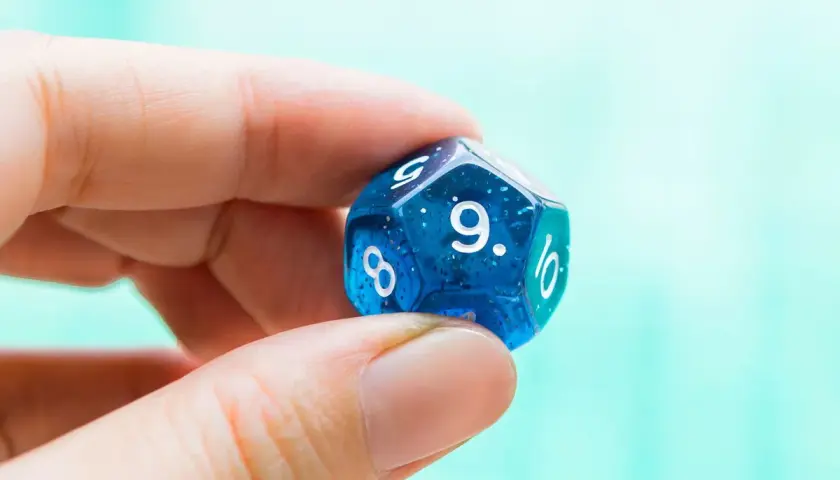 Palec i kciuk trzymają niebieską, dwudziestościenną kostkę do gry z widoczną cyfrą 9. To symbol numerologiczna 9, oznaczająca zakończenie cyklu.